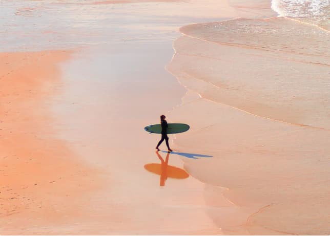 Lone surfer walking across a peach-coloured beach.